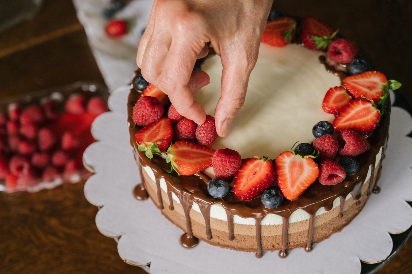 Hand placing fresh strawberries on a chocolate drip cake topped with berries at Crumb and Kiln Bakery